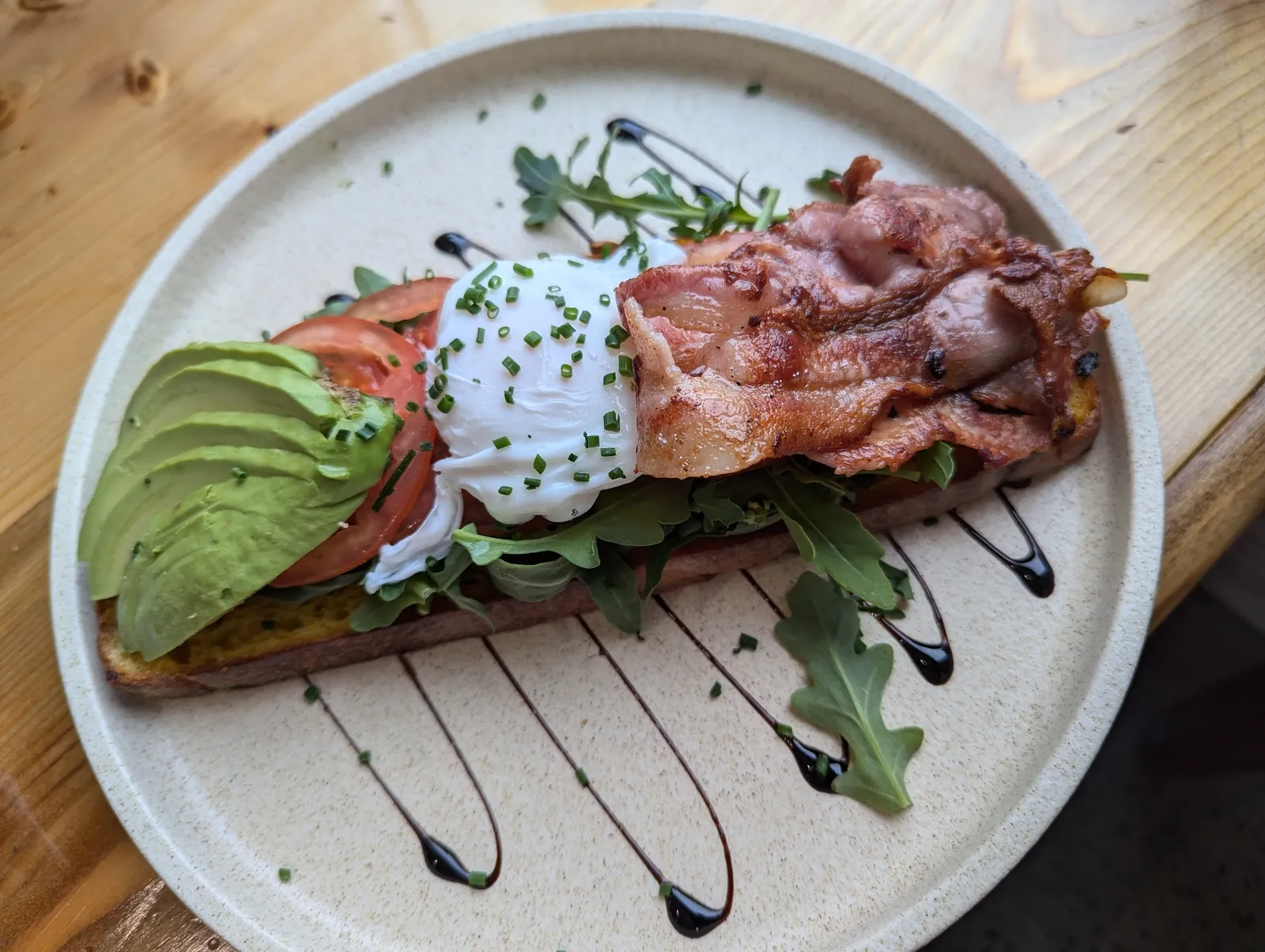 Avocado toast with poached egg, bacon, rocket and balsamic drizzle on cream ceramic plate