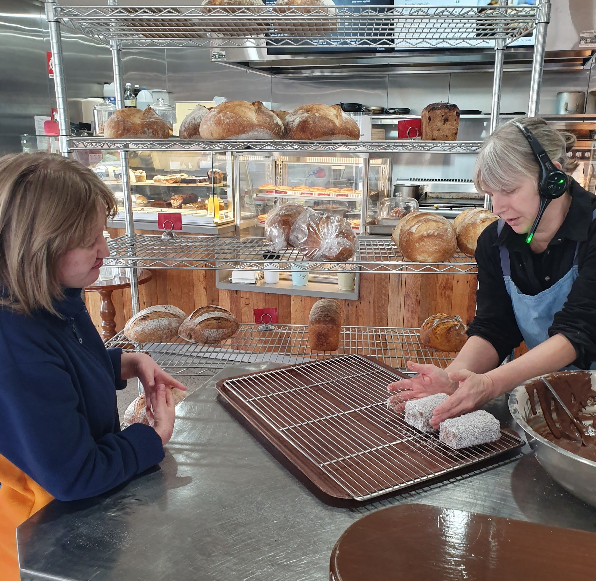 Two Cafe Viana staff preparing lamingtons at the bakery counter with sourdough loaves on shelves behind