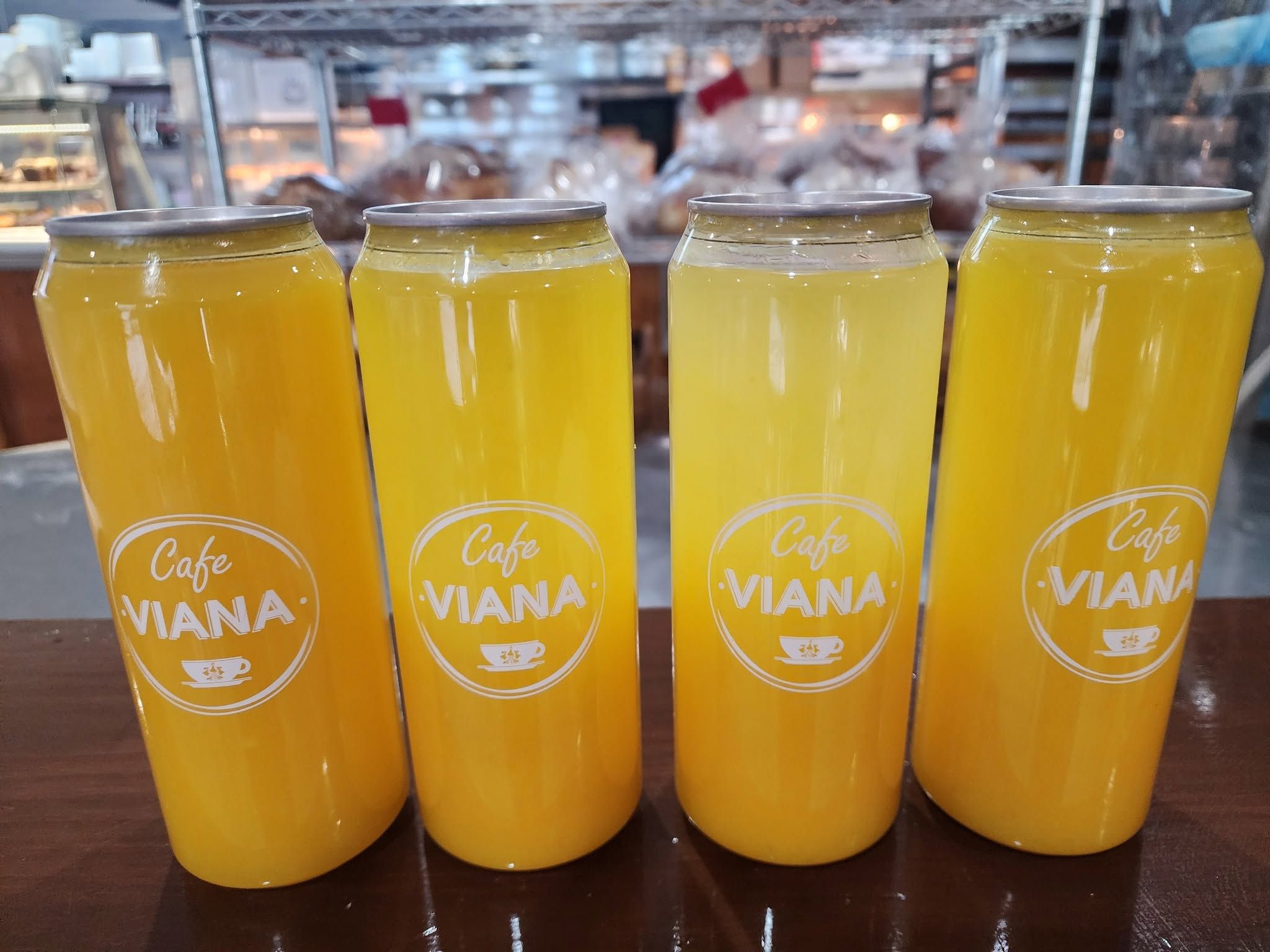 Four Cafe Viana branded yellow juice cans in a row with bakery shelves in the background